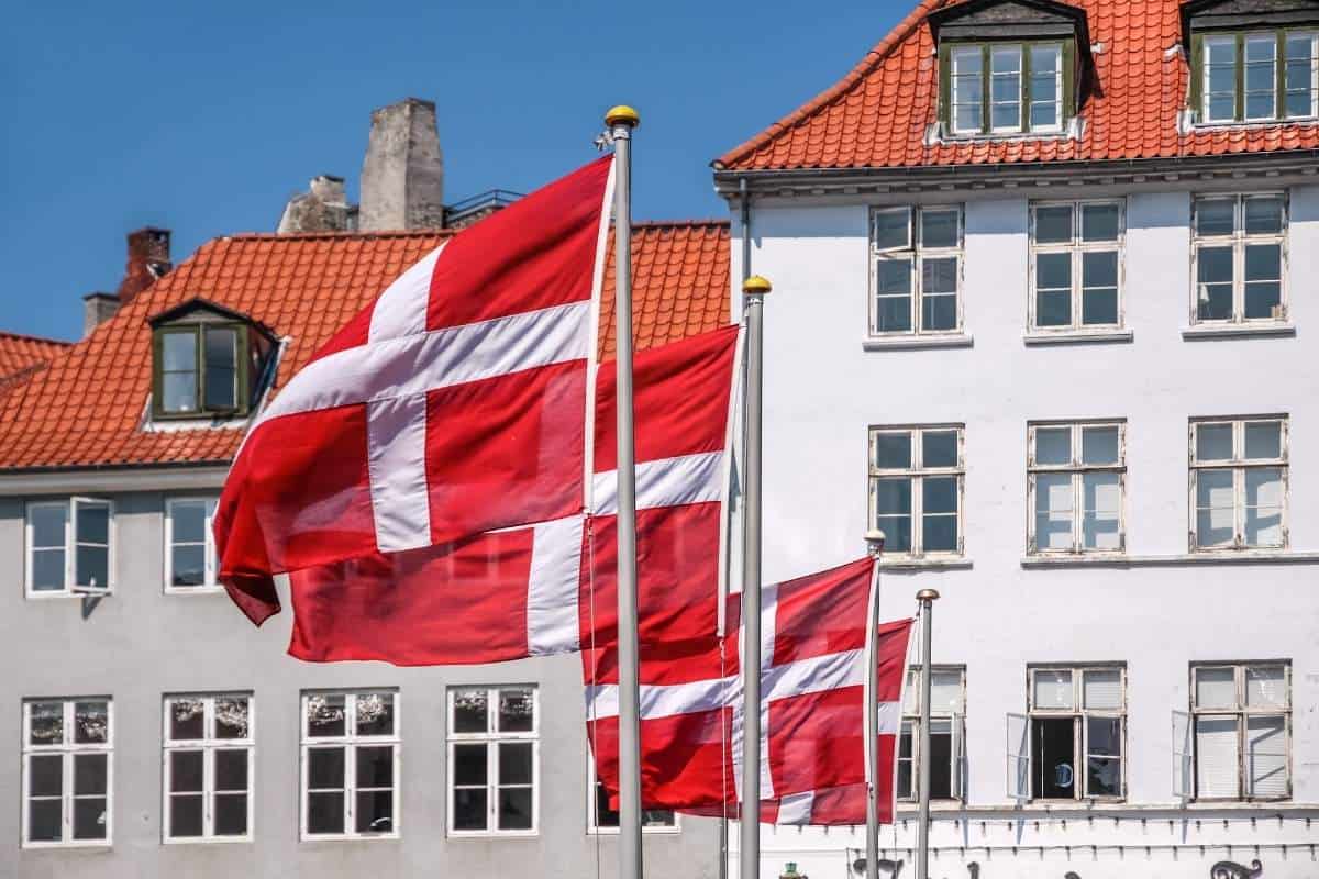A row of Danish flags blowing in the breeze in front of white buildings on a clear day