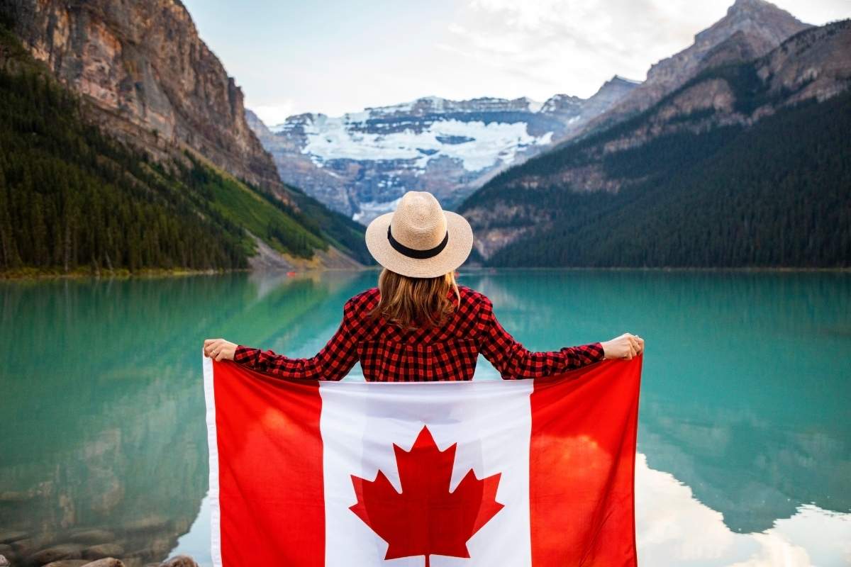 The post about interesting facts about Canada. Woman holding a Canadian flag looking at Lake Louise in Alberta, Canada.