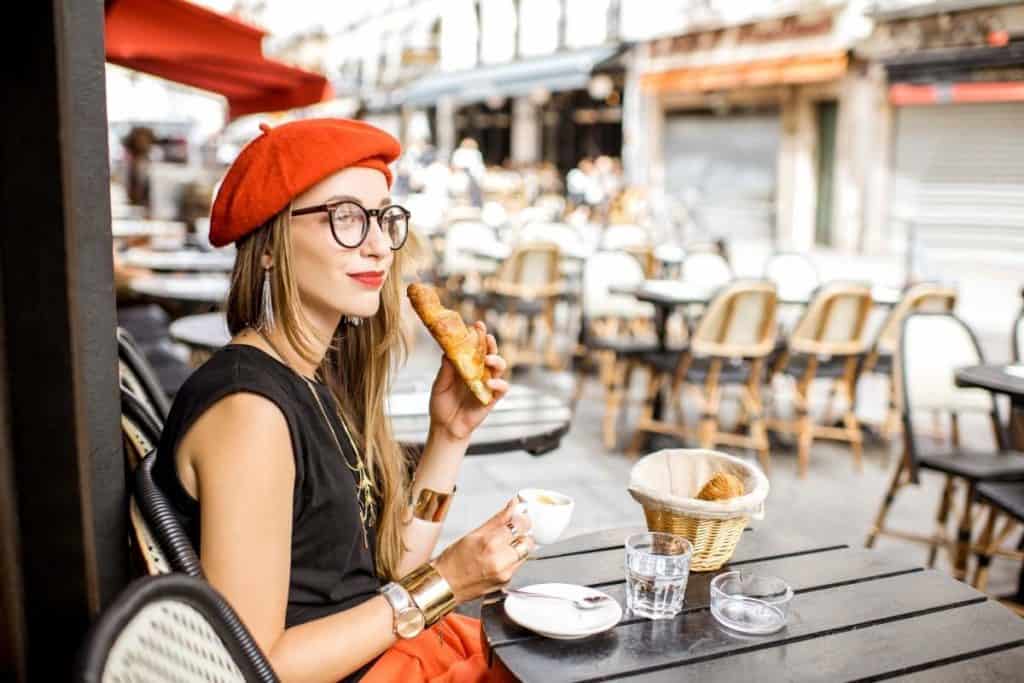 Young stylish woman wearing a red beret having a French breakfast with coffee and croissant at an outdoor patio