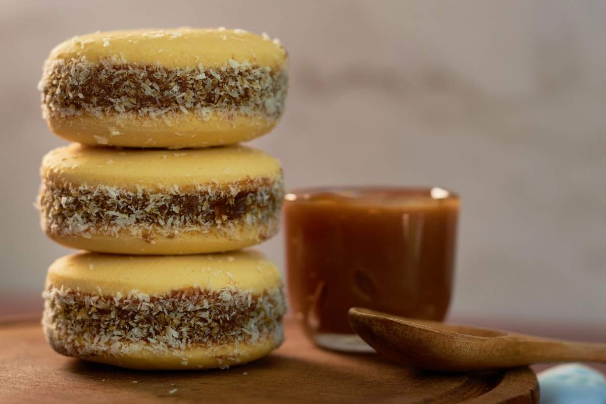 Three stacked alfajores cookies, a dessert from Argentina, with a clear cup of dulce de leche with a wooden spoon on a wooden table