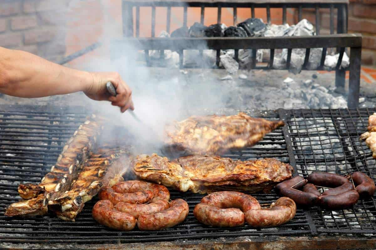 View of a traditional Argentinian asado (bbq)multiple grilled meats cooked over an open flame with smoke rising up and a hand emerging to tend to them
