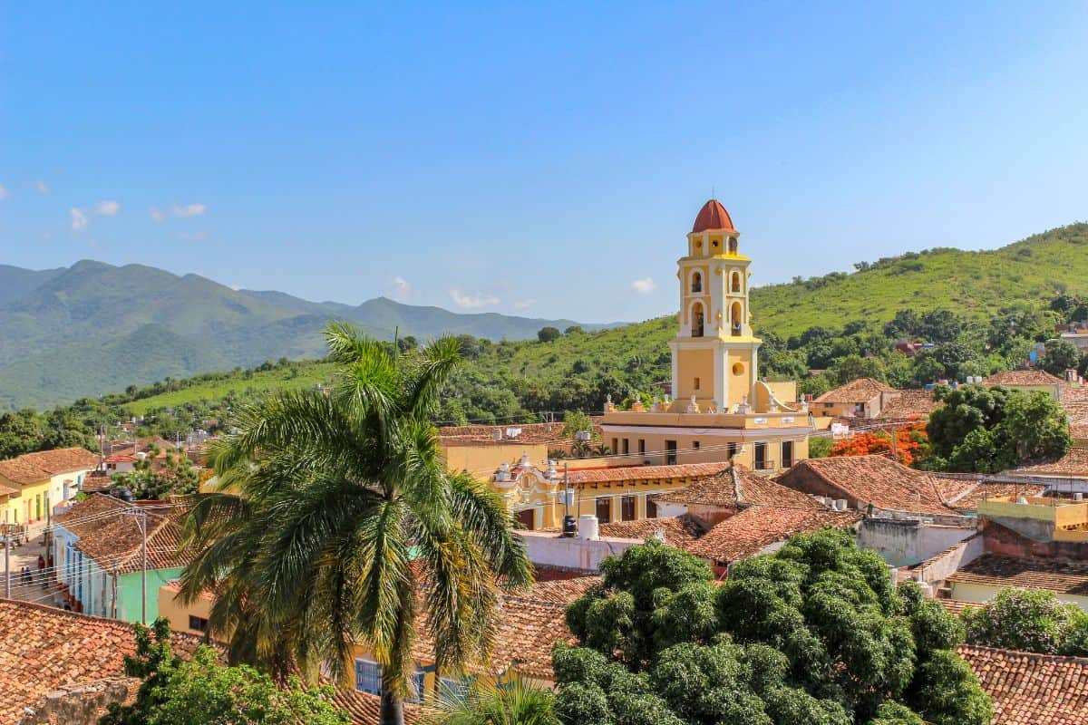 Towering yellow Catholic church with an orange roof surrounded by palm trees and mountainous jungle with a clear blue sky in the background