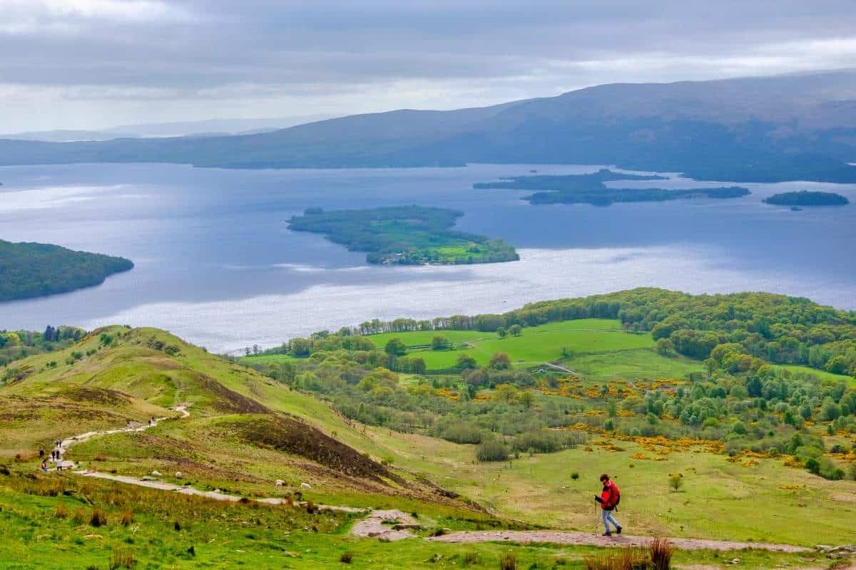 A person wearing a red coat walking a trail within Loch Lomond & The Trossachs National Park in Scotland on a cloudy day with greenery and a large body of water in the background