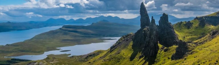 Panoramic view of the Storr, a rocky hill on the Isle of Skye in the Scottish Highlands with a sweeping view of greenery, mountains, and the sea in the background with low cloud cover.