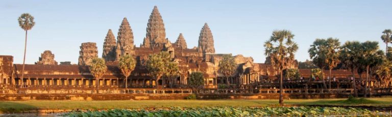 The distinct spires and architecture of Angkor Wat with a clear blue sky behind it and palm trees and green grass in front