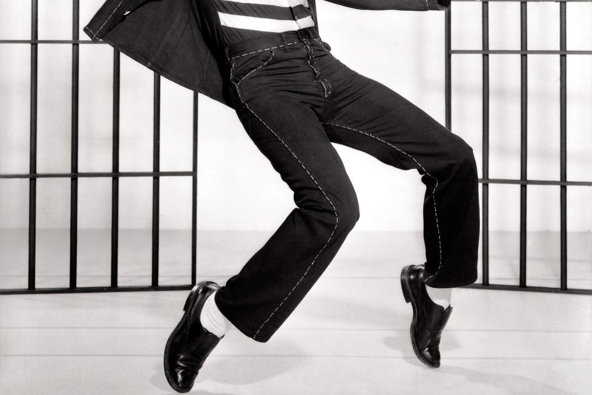 Black and white image of a man from the waist down standing on tip toes with the bars of a jail cell behind him