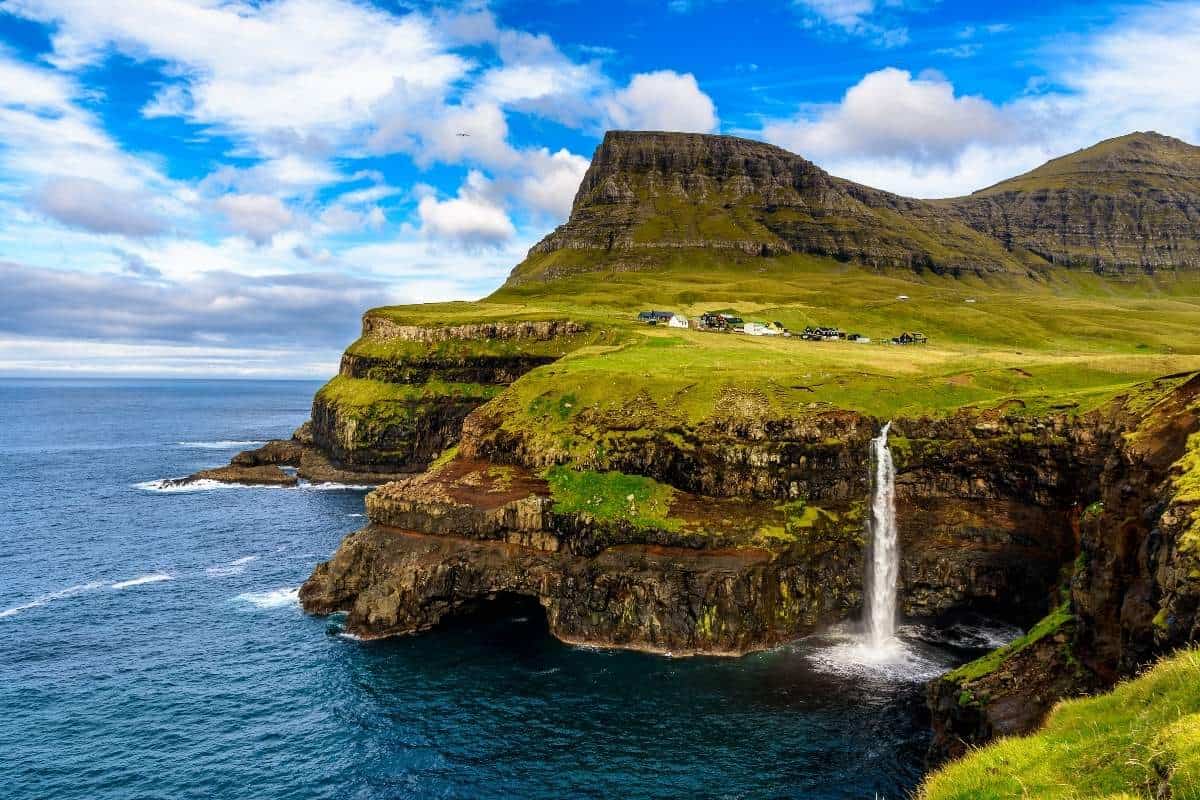 A waterfall flowing over a cliffs edge into the sea with green covering the land and a bright blue sky with fluffy white clouds in the Faroe Islands