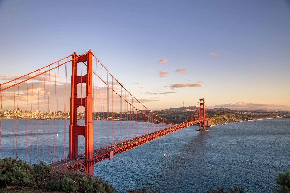 In our list of world famous landmarks we include an aerial image of the red Golden Gate Bridge spanning across open water at sunset with a lone sailboat with a white sail passing by underneath