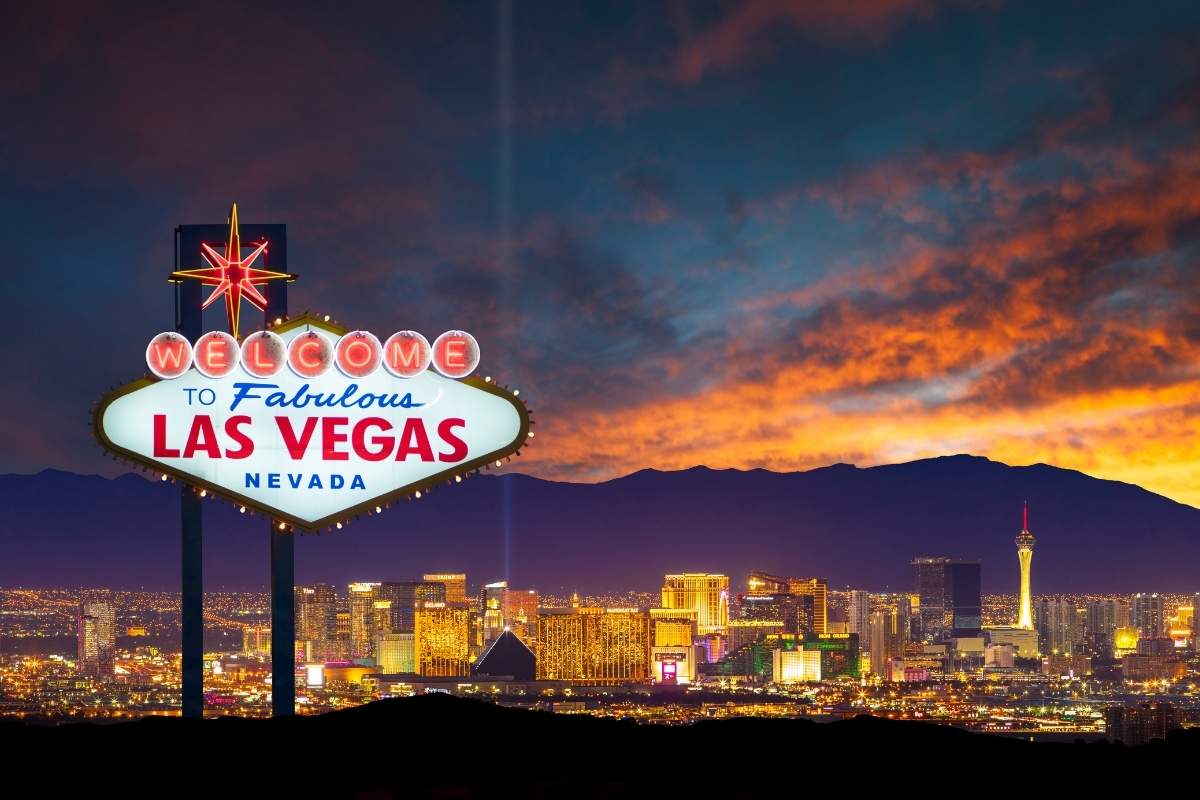 A brightly lit Welcome to Las Vegas sign at sunset with the lights of the Las Vegas Strip in the background