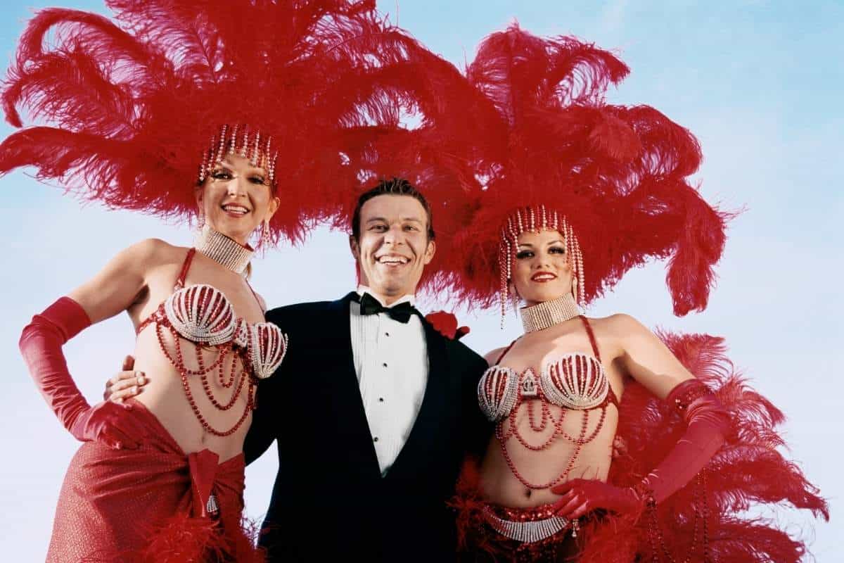Man wearing a tux with his arms around two showgirls dressed in red costumes with feather headdresses