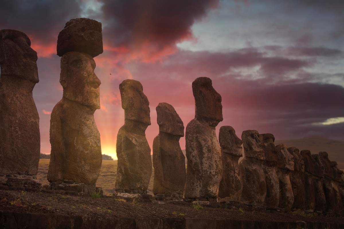 A row of Moais statues from Easter Island lined up in a row with a beautiful pink and purple sunset behind them. Another one of the most famous world landmarks.