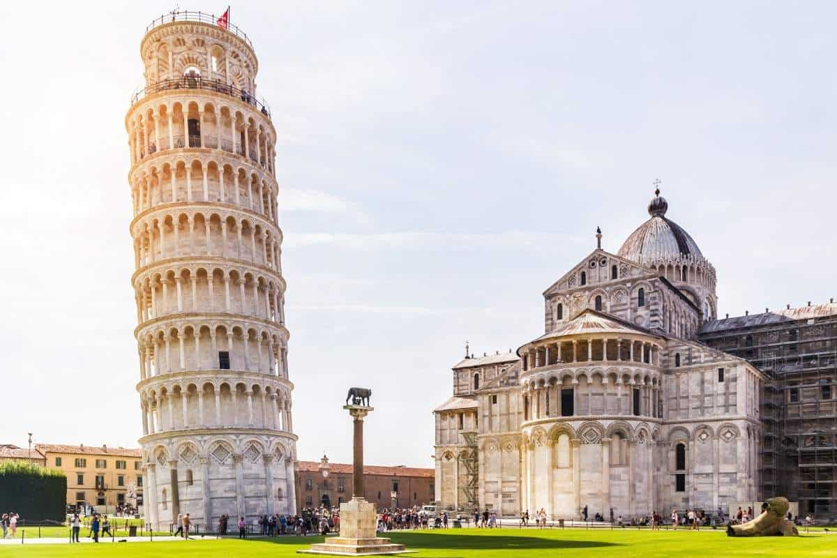 People walking on green grass in front of the Leaning Tower of Pisa and the Pisa Cathedral on a clear, sunny day