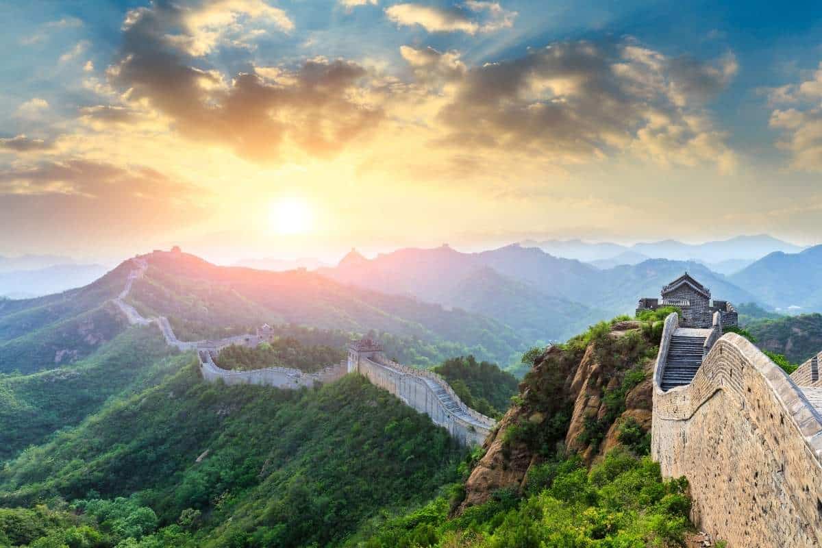 Sunrise behind a cloudy mountain illuminating a stretch of the Great Wall of China with green trees and shrubs alongside it