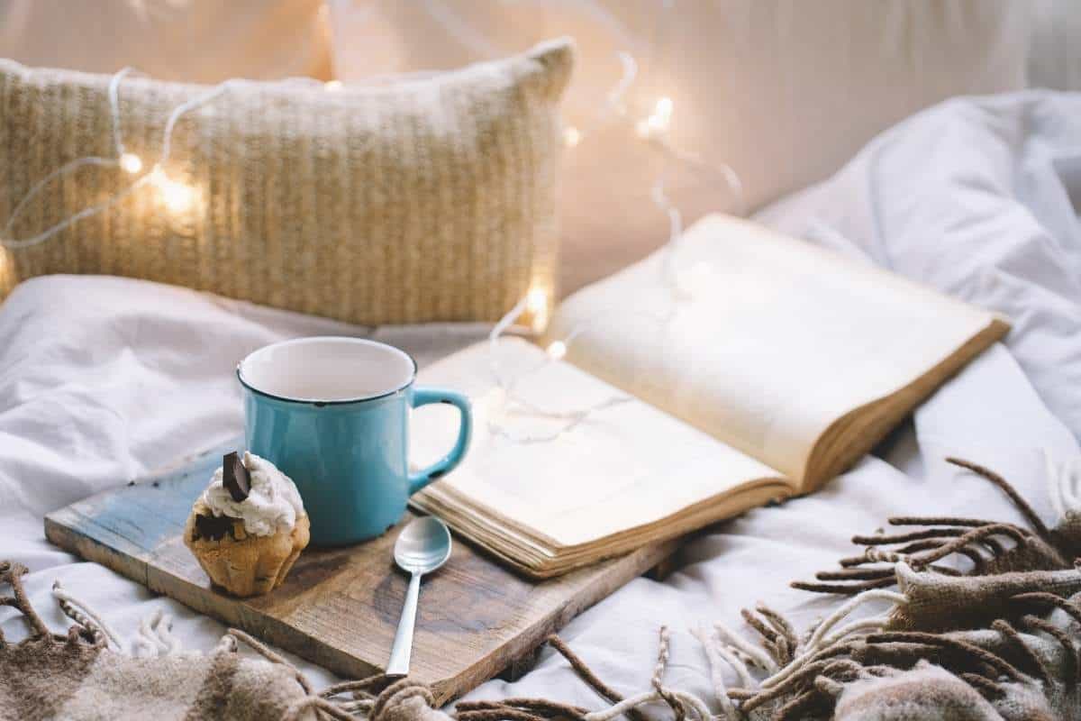 A cozy scene depicting the Danish lifestyle of hygge with an open book, cup of tea and muffin on a wooden board on a bed with twinkle lights and a white plush pillow in the background