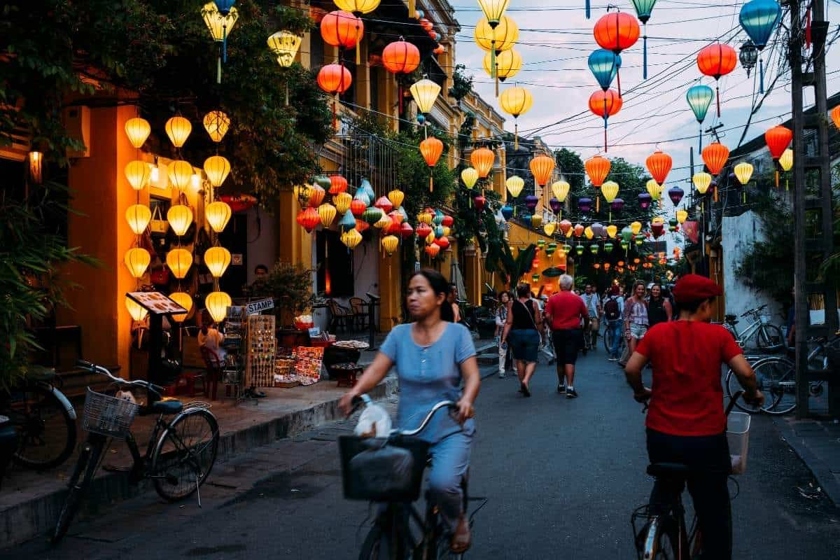 A busy pedestrian street scene in Vietnam at dusk with colorful glowing lanterns hanging overtop and hanging strewn along the street