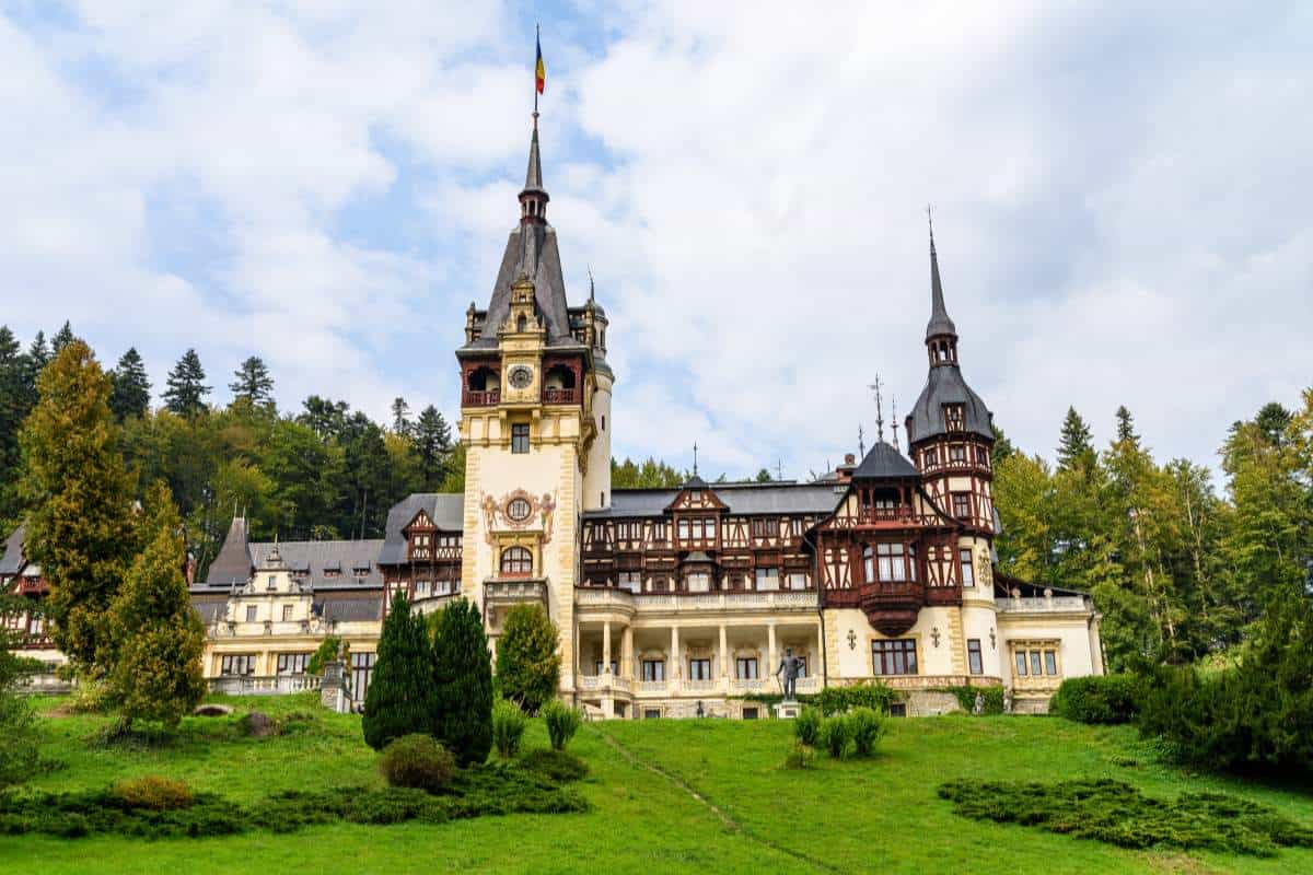 Neo-Renaissance Pele Castle in Romania surrounded by thick green grass and forest amongst a blue sky with white fluffy clouds