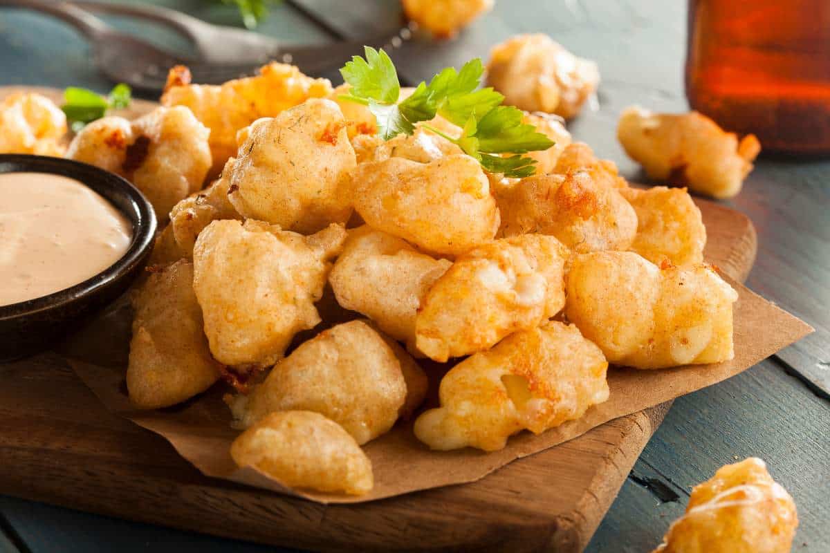 a pile of deep fried cheese curds topped with green herbs sitting on a wooden board with a side of dipping sauce. 60+ Facts about US states