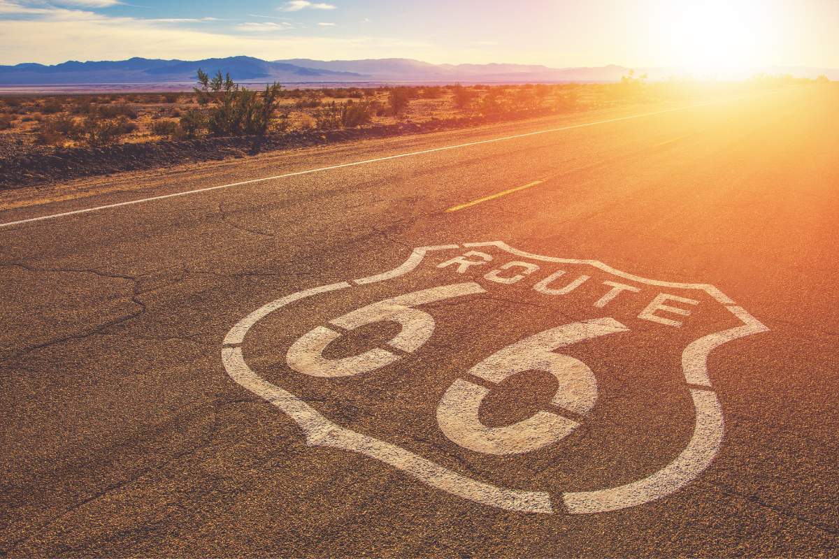 Close up perspective of a paved road with Route 66 painted in white with the sun coming in at an angle at the top right and a desert landscape behind it