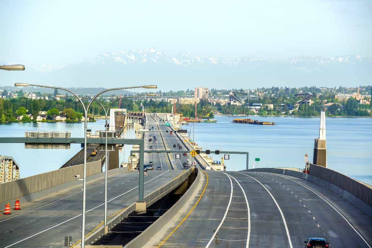 Road perspective of Evergreen Point floating bridge in Washington, USA going over water with a cityscape and mountains in the distance. 60+ Facts about US states