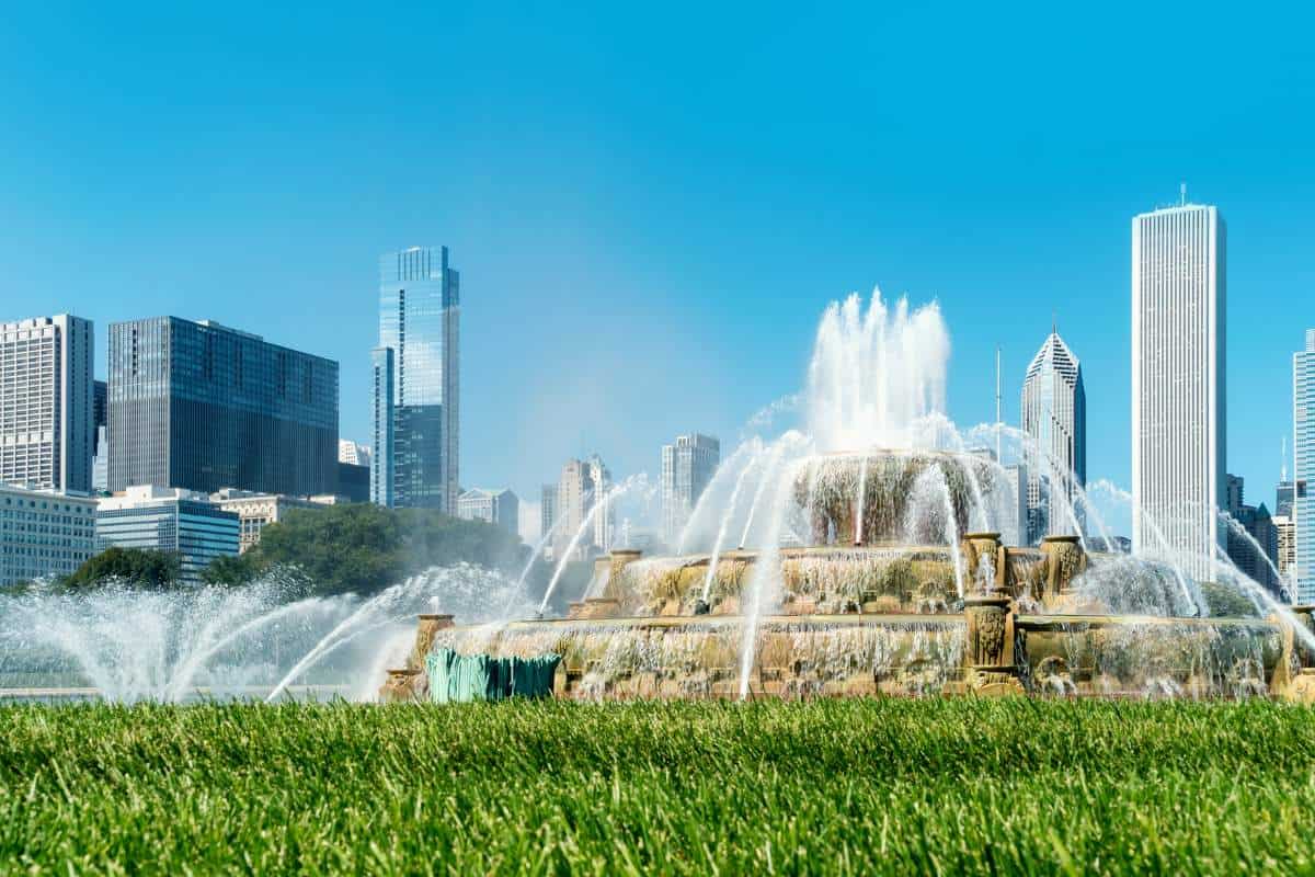 Close up view of a water fountain in action on a clear sunny day with green grass in the foreground and a cityscape behind