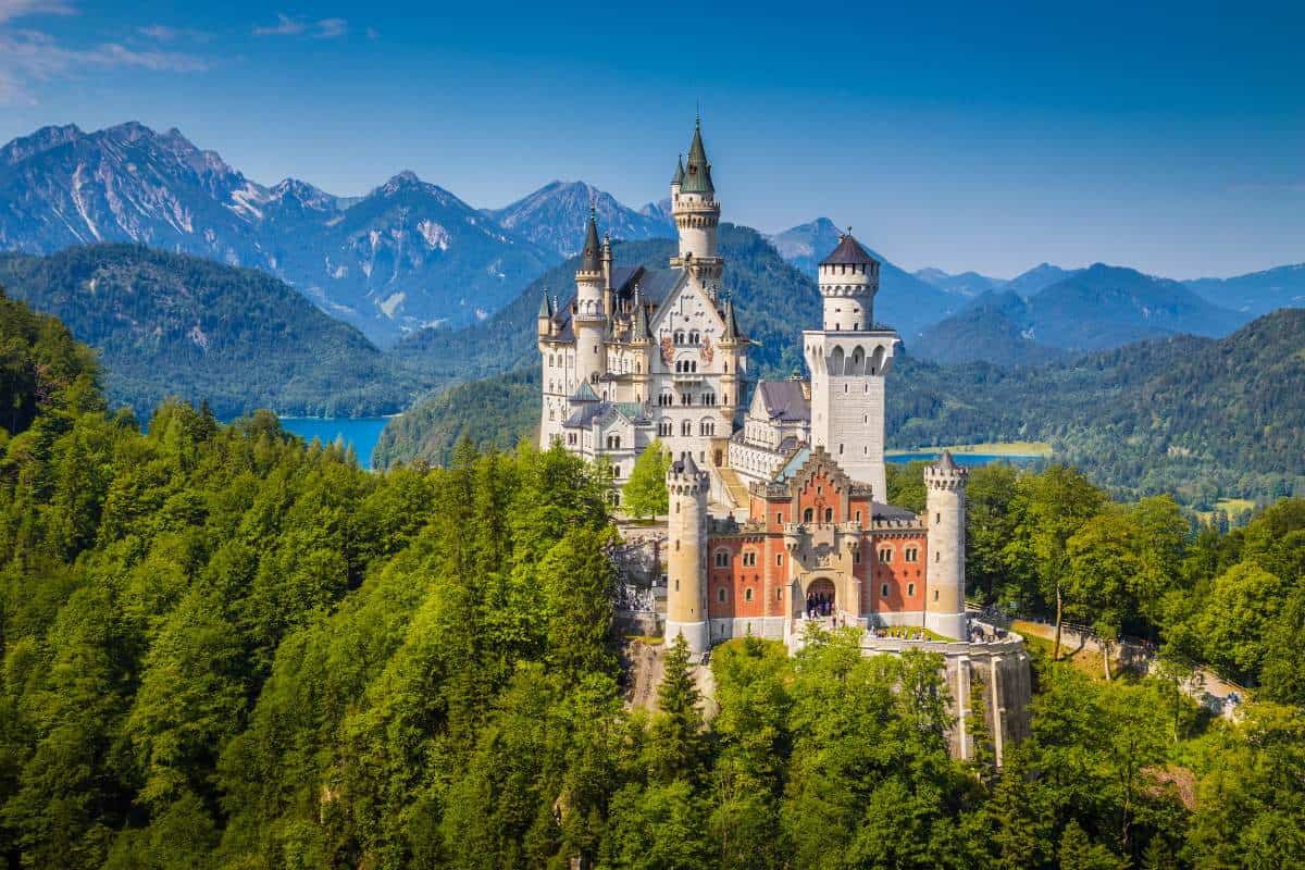 Romantic castles with multiple turrets sitting amidst and on top green trees with mountains and a blue lake visible in the distance