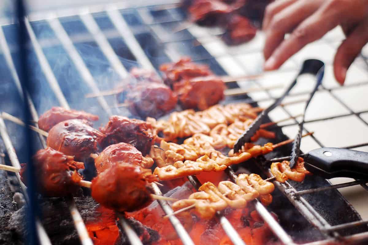 Skewered meat and Filipino delicacies on a grill with a hand holding tongs reaching for them