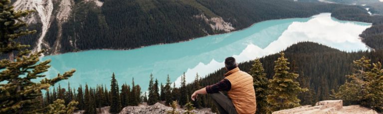 Man wearing a yellow vest sitting by himself on a rock ledge looking down at the blue water of a glacier lake in the Canadian Rocky Mountains